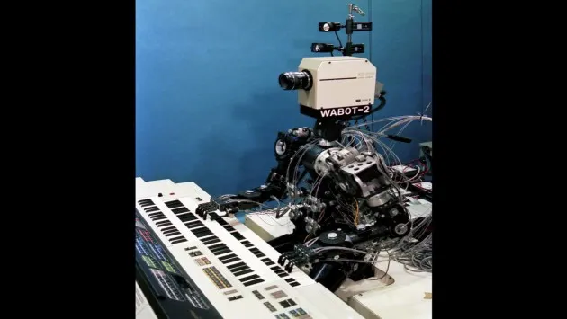 A humanoid robot seated at an electronic organ keyboard, with hands positioned over the keys